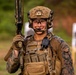 A smiling military service member in full combat gear stands at what appears to be a shooting range. The individual is wearing MARPAT (Marine pattern) camouflage uniform, a tactical helmet with mounted equipment, and a plate carrier vest, holding a rifle upright. Shooting targets are visible in the blurred background.