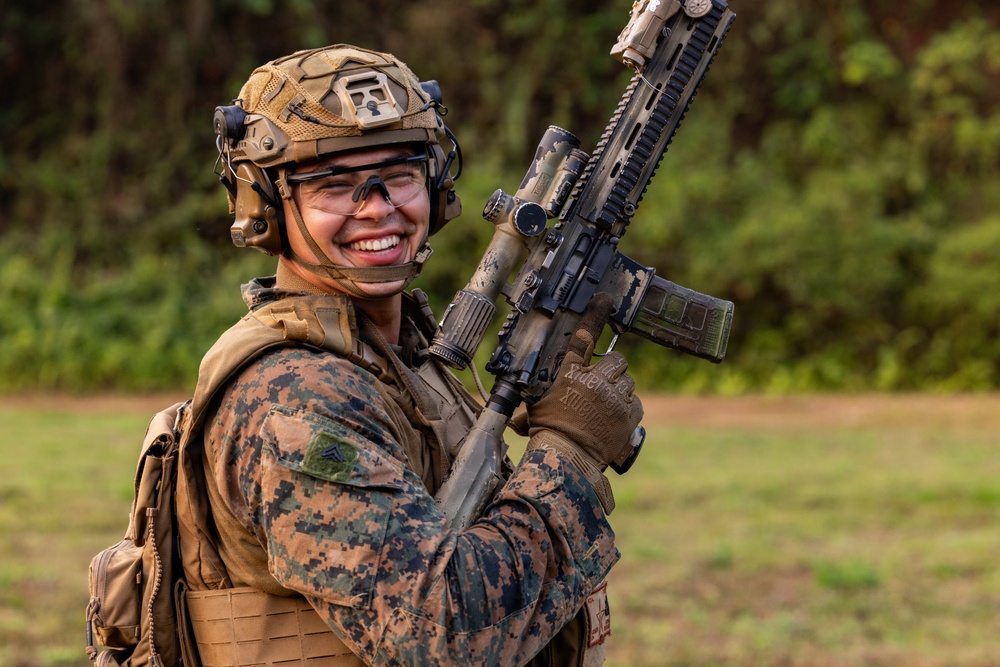 A smiling military service member in full combat gear holds an assault rifle in an outdoor field setting. The individual is wearing USMC digital camouflage (MARPAT), a tactical helmet, body armor, and protective eyewear. The mood appears relaxed and positive despite the full combat loadout.