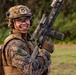 A smiling military service member in full combat gear holds an assault rifle in an outdoor field setting. The individual is wearing USMC digital camouflage (MARPAT), a tactical helmet, body armor, and protective eyewear. The mood appears relaxed and positive despite the full combat loadout.