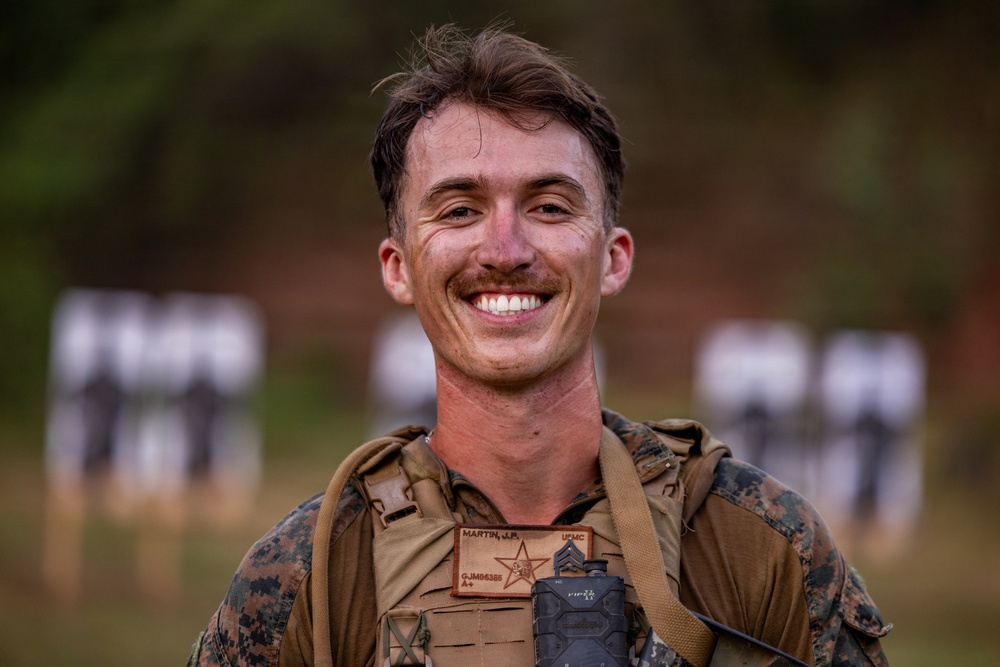 A smiling male service member wearing USMC (United States Marine Corps) digital camouflage uniform and a tan tactical plate carrier vest is photographed at what appears to be a shooting range. His name tape reads 'MARTIN, J.R.' and the branch tape reads 'USMC'. Blurred silhouette targets are visible in the background.