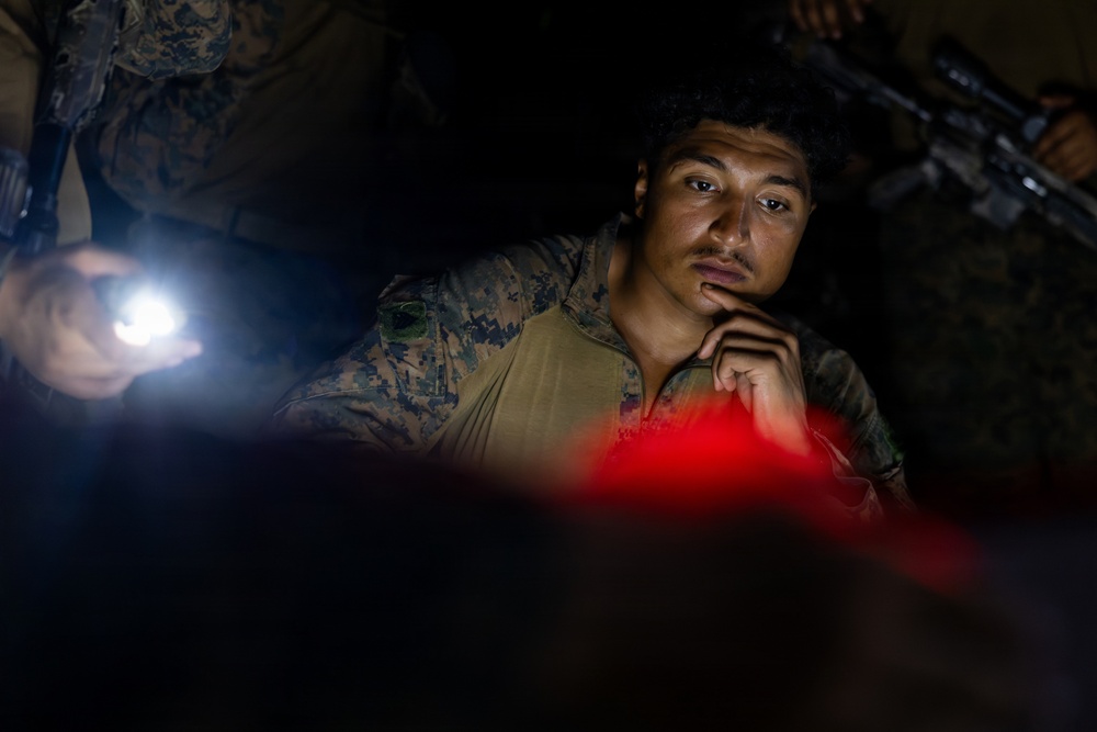 A military personnel wearing MARPAT (Marine pattern) camouflage uniform is visible in a low-light environment, illuminated by a flashlight and a red light source. The individual appears to be in a briefing or planning session, with their hand raised to their chin in a contemplative pose. Other armed personnel are partially visible in the background.