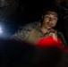 A military personnel wearing MARPAT (Marine pattern) camouflage uniform is visible in a low-light environment, illuminated by a flashlight and a red light source. The individual appears to be in a briefing or planning session, with their hand raised to their chin in a contemplative pose. Other armed personnel are partially visible in the background.