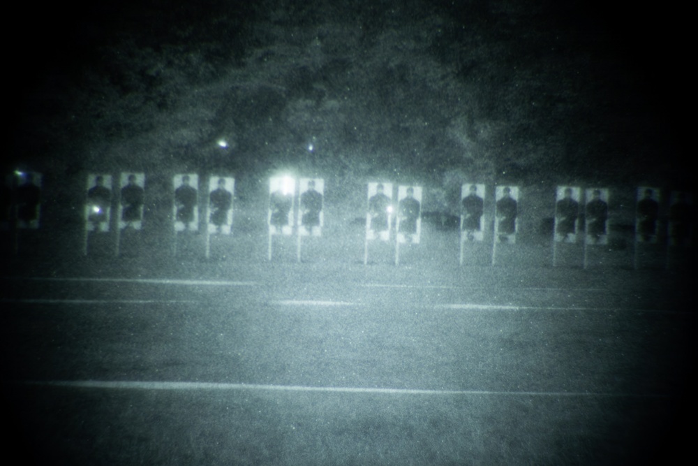 A night-vision or low-light photograph showing a row of human silhouette shooting targets arranged on a range or open field. The image appears to be taken through a night-vision device or with a low-light camera, giving it a characteristic green-gray grainy appearance. Several bright light flares are visible among the targets, suggesting active shooting or illumination sources.