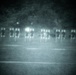 A night-vision or low-light photograph showing a row of human silhouette shooting targets arranged on a range or open field. The image appears to be taken through a night-vision device or with a low-light camera, giving it a characteristic green-gray grainy appearance. Several bright light flares are visible among the targets, suggesting active shooting or illumination sources.