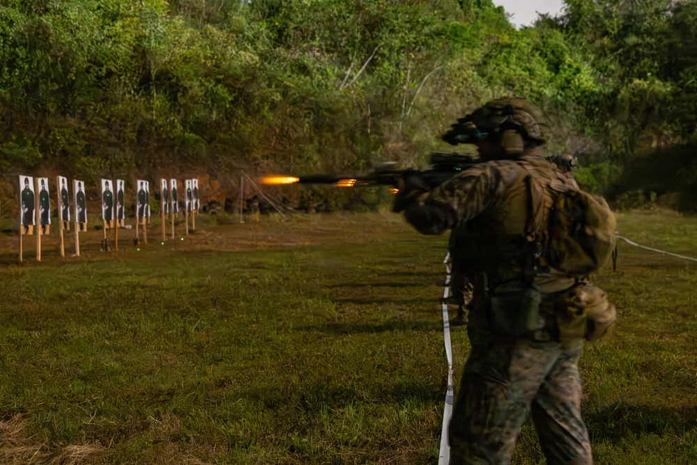 A fully equipped military or law enforcement operator in camouflage gear is firing a rifle at a live-fire shooting range, with a visible muzzle flash captured mid-shot. Multiple paper silhouette targets are lined up downrange against a natural earthen berm backdrop. The setting appears to be an outdoor training range surrounded by dense tropical or subtropical vegetation.