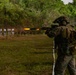 A fully equipped military or law enforcement operator in camouflage gear is firing a rifle at a live-fire shooting range, with a visible muzzle flash captured mid-shot. Multiple paper silhouette targets are lined up downrange against a natural earthen berm backdrop. The setting appears to be an outdoor training range surrounded by dense tropical or subtropical vegetation.