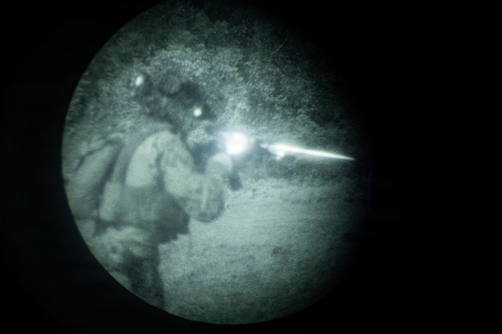 This image is captured through a night vision device, showing a circular field of view with characteristic green-gray monochrome tones. A figure in tactical/military gear appears to be firing a weapon, as evidenced by a bright muzzle flash visible at the center-right of the frame. The image is taken during nighttime conditions.