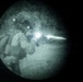 This image is captured through a night vision device, showing a circular field of view with characteristic green-gray monochrome tones. A figure in tactical/military gear appears to be firing a weapon, as evidenced by a bright muzzle flash visible at the center-right of the frame. The image is taken during nighttime conditions.