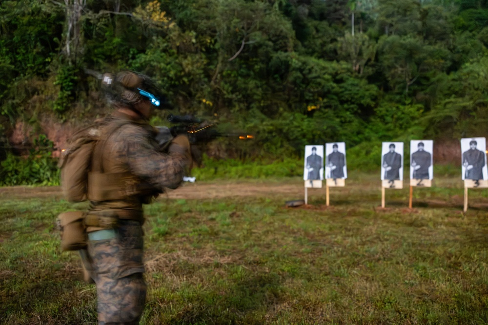 A military or law enforcement personnel in camouflage gear is firing a rifle at a shooting range, with visible muzzle flash captured in the image. Five human-silhouette paper targets are mounted on wooden stakes downrange. The shooter appears to be in motion, causing motion blur in the photograph.