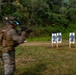 A military or law enforcement personnel in camouflage gear is firing a rifle at a shooting range, with visible muzzle flash captured in the image. Five human-silhouette paper targets are mounted on wooden stakes downrange. The shooter appears to be in motion, causing motion blur in the photograph.