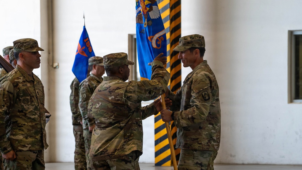 A military change of command or guidon ceremony is taking place inside what appears to be a hangar or large military building. A central figure in OCP (Operational Camouflage Pattern) uniform is passing or receiving a unit guidon/flag to another soldier, a formal ritual signifying transfer of command authority. Multiple soldiers in matching camouflage uniforms are standing in formation as witnesses to the ceremony.