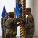 A military change of command or guidon ceremony is taking place inside what appears to be a hangar or large military building. A central figure in OCP (Operational Camouflage Pattern) uniform is passing or receiving a unit guidon/flag to another soldier, a formal ritual signifying transfer of command authority. Multiple soldiers in matching camouflage uniforms are standing in formation as witnesses to the ceremony.