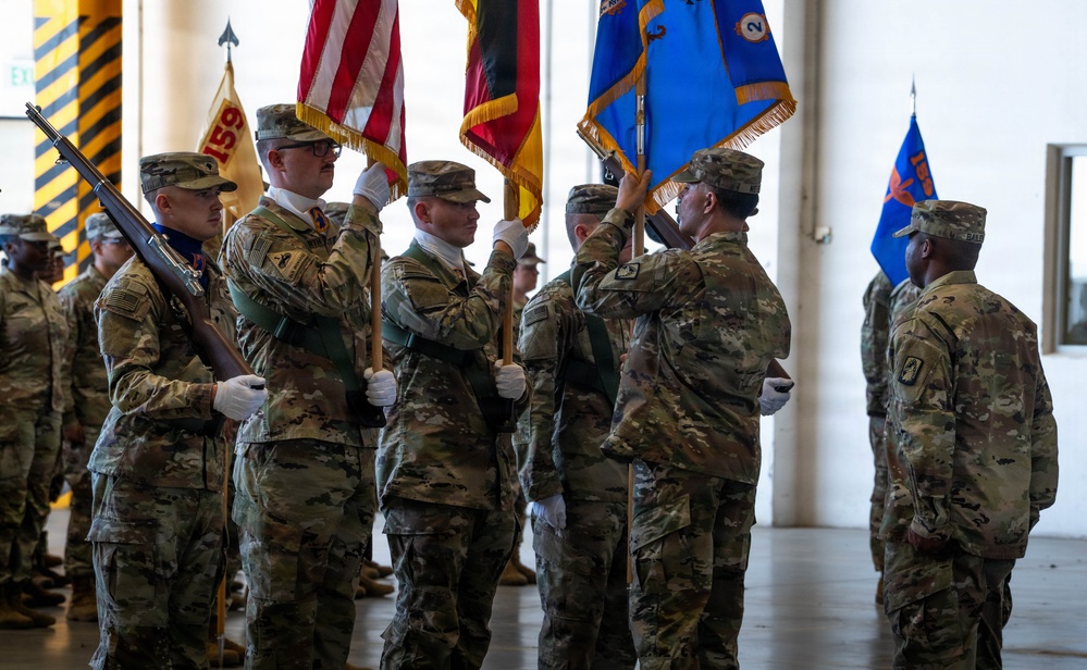 A U.S. Army military ceremony, likely a change of command or activation/inactivation ceremony, taking place inside a large indoor facility. Soldiers in OCP (Operational Camouflage Pattern) uniforms are participating in a color guard formation, presenting and transferring unit flags and guidons. Multiple flags are visible including the U.S. national flag and unit colors with gold fringe.
