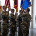 A U.S. Army military ceremony, likely a change of command or activation/inactivation ceremony, taking place inside a large indoor facility. Soldiers in OCP (Operational Camouflage Pattern) uniforms are participating in a color guard formation, presenting and transferring unit flags and guidons. Multiple flags are visible including the U.S. national flag and unit colors with gold fringe.