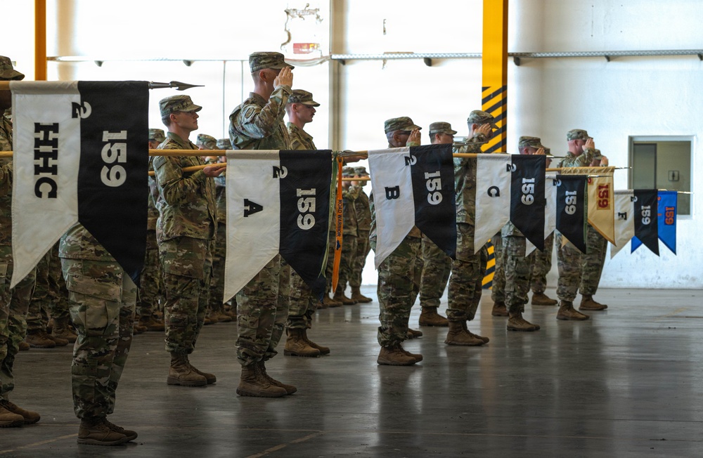 A formal U.S. Army military ceremony taking place inside what appears to be a hangar or large indoor facility. Soldiers in OCP (Operational Camouflage Pattern) uniforms are standing in formation holding guidon pennants bearing the number '159' and various company designations including HHC, A, B, C, and E. Several soldiers in the background are rendering salutes, indicating an official ceremony such as a change of command, activation, or deployment.