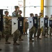 A formal U.S. Army military ceremony taking place inside what appears to be a hangar or large indoor facility. Soldiers in OCP (Operational Camouflage Pattern) uniforms are standing in formation holding guidon pennants bearing the number '159' and various company designations including HHC, A, B, C, and E. Several soldiers in the background are rendering salutes, indicating an official ceremony such as a change of command, activation, or deployment.