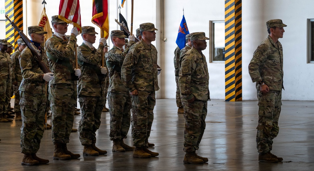 A formal military ceremony is taking place inside what appears to be a large hangar or indoor facility. Soldiers in US Army OCP (Operational Camouflage Pattern) uniforms are standing at attention, with a color guard holding multiple flags including the US flag and unit guidons. The setting and formation suggest this is a change of command, activation, or similar official military ceremony.