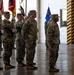 A formal military ceremony is taking place inside what appears to be a large hangar or indoor facility. Soldiers in US Army OCP (Operational Camouflage Pattern) uniforms are standing at attention, with a color guard holding multiple flags including the US flag and unit guidons. The setting and formation suggest this is a change of command, activation, or similar official military ceremony.