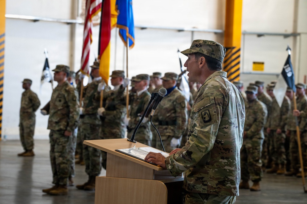 A military officer in U.S. Army camouflage (OCP) uniform is delivering a speech at a podium during a formal military ceremony held inside a large hangar or indoor facility. Behind the speaker, multiple soldiers stand in formation holding color guard flags, including the U.S. flag and what appears to be a German flag, suggesting a multinational or NATO-related event. The setting and arrangement indicate a change of command, activation, or unit ceremony.