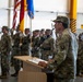 A military officer in U.S. Army camouflage (OCP) uniform is delivering a speech at a podium during a formal military ceremony held inside a large hangar or indoor facility. Behind the speaker, multiple soldiers stand in formation holding color guard flags, including the U.S. flag and what appears to be a German flag, suggesting a multinational or NATO-related event. The setting and arrangement indicate a change of command, activation, or unit ceremony.