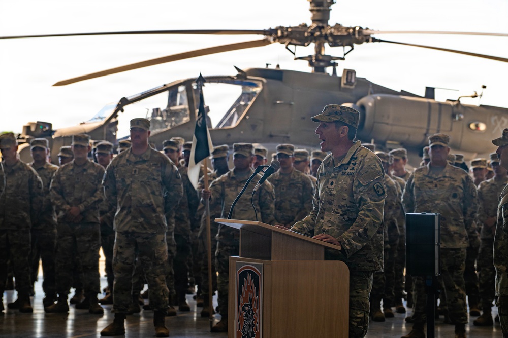 A U.S. Army officer is delivering a speech at a podium during a military ceremony held inside an aircraft hangar. A large formation of soldiers in MultiCam/OCP camouflage uniforms stands at attention in the background. An Apache attack helicopter is prominently visible behind the assembled troops.