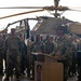 A U.S. Army officer is delivering a speech at a podium during a military ceremony held inside an aircraft hangar. A large formation of soldiers in MultiCam/OCP camouflage uniforms stands at attention in the background. An Apache attack helicopter is prominently visible behind the assembled troops.