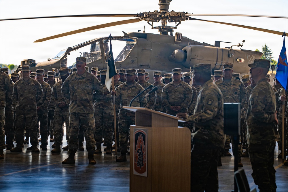 A formal military ceremony is taking place inside what appears to be a hangar or large indoor facility. A uniformed soldier is addressing a large formation of troops from a wooden podium, with an AH-64 Apache attack helicopter serving as a backdrop. The event appears to be a change of command, unit activation, or similar official military ceremony.