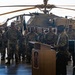 A formal military ceremony is taking place inside what appears to be a hangar or large indoor facility. A uniformed soldier is addressing a large formation of troops from a wooden podium, with an AH-64 Apache attack helicopter serving as a backdrop. The event appears to be a change of command, unit activation, or similar official military ceremony.