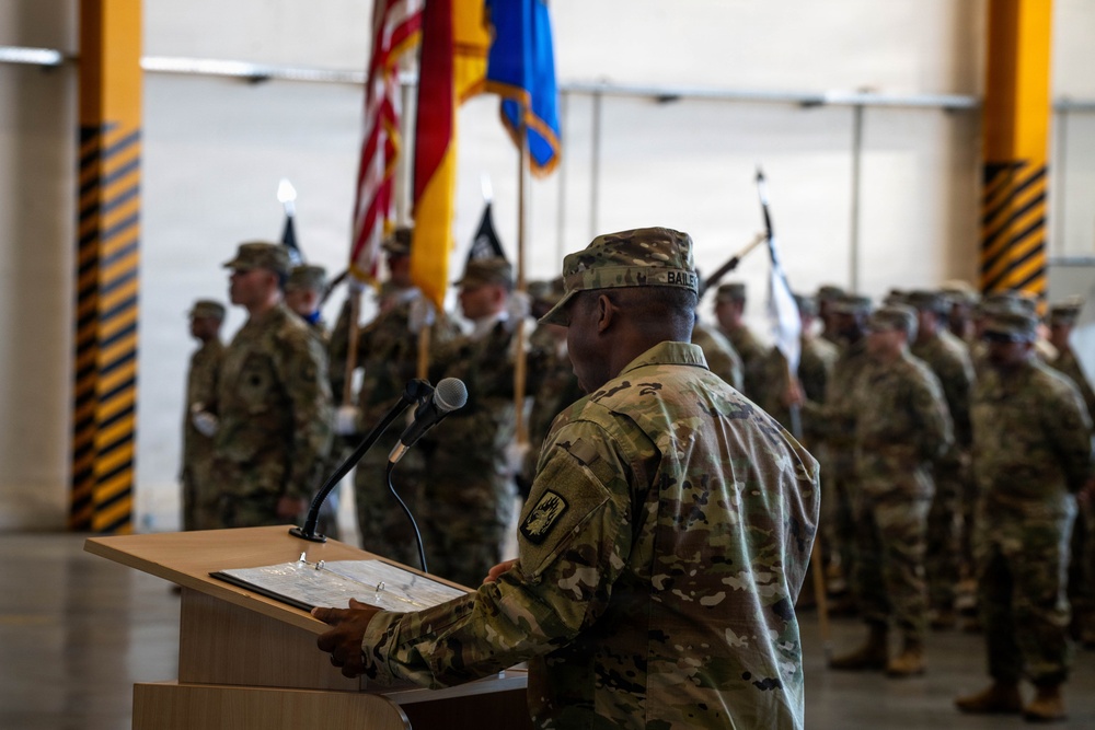 A military ceremony is taking place inside what appears to be a hangar or large indoor facility. A soldier identified by nametape as 'BAILEY' stands at a wooden podium addressing a formation of uniformed soldiers in the background. Multiple flags, including the U.S. flag and unit colors, are visible being held by a color guard.