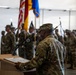 A military ceremony is taking place inside what appears to be a hangar or large indoor facility. A soldier identified by nametape as 'BAILEY' stands at a wooden podium addressing a formation of uniformed soldiers in the background. Multiple flags, including the U.S. flag and unit colors, are visible being held by a color guard.