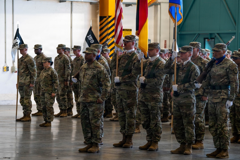 A U.S. Army military formation ceremony is taking place inside what appears to be a large hangar or indoor facility. Soldiers in Army Combat Uniforms (ACU/OCP camouflage pattern) are standing at attention in organized ranks, with a color guard prominently displaying multiple flags including the U.S. flag, a German flag, and unit guidons. The event appears to be a formal military ceremony such as a change of command, activation, or unit formation.