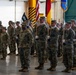 A U.S. Army military formation ceremony is taking place inside what appears to be a large hangar or indoor facility. Soldiers in Army Combat Uniforms (ACU/OCP camouflage pattern) are standing at attention in organized ranks, with a color guard prominently displaying multiple flags including the U.S. flag, a German flag, and unit guidons. The event appears to be a formal military ceremony such as a change of command, activation, or unit formation.