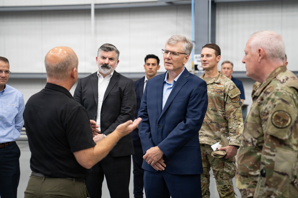 A group of individuals consisting of both civilians and military personnel in camouflage uniforms are gathered inside what appears to be a large industrial or hangar facility. A bald man in a black polo shirt appears to be briefing or presenting to the group, using hand gestures. The attendees are listening attentively in a semi-circle formation.