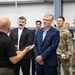 A group of individuals consisting of both civilians and military personnel in camouflage uniforms are gathered inside what appears to be a large industrial or hangar facility. A bald man in a black polo shirt appears to be briefing or presenting to the group, using hand gestures. The attendees are listening attentively in a semi-circle formation.