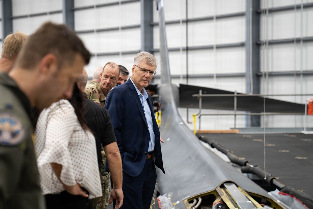 A group of individuals, including military personnel in camouflage uniforms and civilians in business attire, are gathered inside a large industrial hangar examining aircraft components. The central figure, a man in a navy blue suit with glasses, appears to be closely inspecting what appears to be a disassembled or partially assembled aircraft structure. The setting suggests a formal facility tour or technical briefing at an aerospace or defense installation.