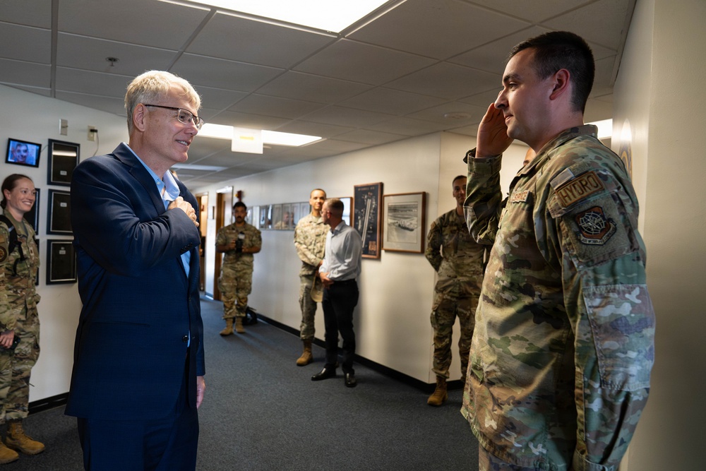 A civilian in a navy blue suit is interacting with a military service member in OCP (Operational Camouflage Pattern) uniform who is rendering a salute. The setting appears to be an indoor military facility hallway, with several other uniformed personnel and at least one other civilian present in the background. The service member's shoulder patch reads 'HYDRO' and displays what appears to be a U.S. Air Force or Space Force unit insignia.