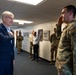 A civilian in a navy blue suit is interacting with a military service member in OCP (Operational Camouflage Pattern) uniform who is rendering a salute. The setting appears to be an indoor military facility hallway, with several other uniformed personnel and at least one other civilian present in the background. The service member's shoulder patch reads 'HYDRO' and displays what appears to be a U.S. Air Force or Space Force unit insignia.