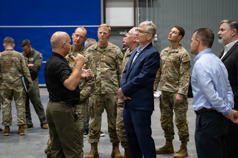 A group of U.S. Air Force personnel in OCP (Operational Camouflage Pattern) uniforms are gathered inside what appears to be a military hangar or large facility. A civilian in a navy blue suit and a man in a black polo shirt appear to be engaged in a briefing or discussion with the military members. The setting suggests an official visit, inspection, or operational briefing.