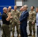A group of U.S. Air Force personnel in OCP (Operational Camouflage Pattern) uniforms are gathered inside what appears to be a military hangar or large facility. A civilian in a navy blue suit and a man in a black polo shirt appear to be engaged in a briefing or discussion with the military members. The setting suggests an official visit, inspection, or operational briefing.
