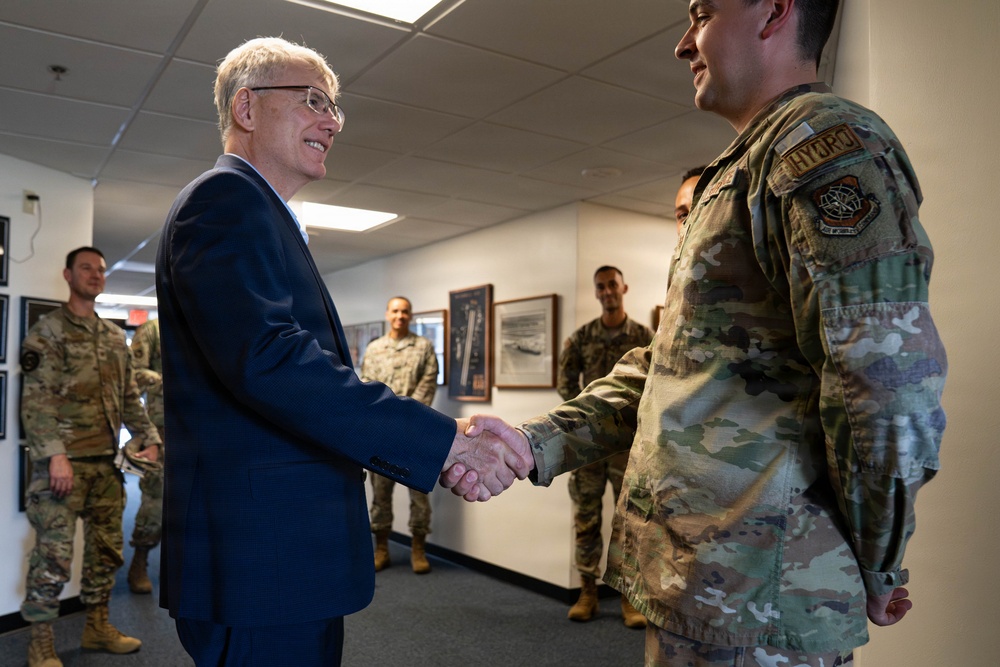 A civilian in a dark blue suit is shaking hands with a military service member wearing OCP (Operational Camouflage Pattern) uniform in what appears to be an official meeting or visit. Several other uniformed military personnel are visible in the background observing the interaction. The setting appears to be an indoor military facility or office building.