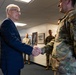 A civilian in a dark blue suit is shaking hands with a military service member wearing OCP (Operational Camouflage Pattern) uniform in what appears to be an official meeting or visit. Several other uniformed military personnel are visible in the background observing the interaction. The setting appears to be an indoor military facility or office building.