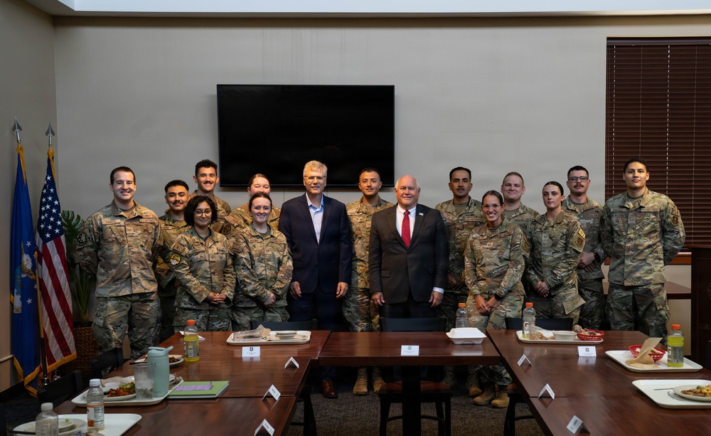 A group photo of approximately 14 military personnel in camouflage uniforms (OCP pattern) posing with two civilians in business attire inside what appears to be a conference or dining room on a military installation. An American flag and what appears to be an Air Force flag are visible in the background. The setting suggests an official visit or meeting, with a dining table set with food trays and water bottles in the foreground.