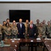 A group photo of approximately 14 military personnel in camouflage uniforms (OCP pattern) posing with two civilians in business attire inside what appears to be a conference or dining room on a military installation. An American flag and what appears to be an Air Force flag are visible in the background. The setting suggests an official visit or meeting, with a dining table set with food trays and water bottles in the foreground.