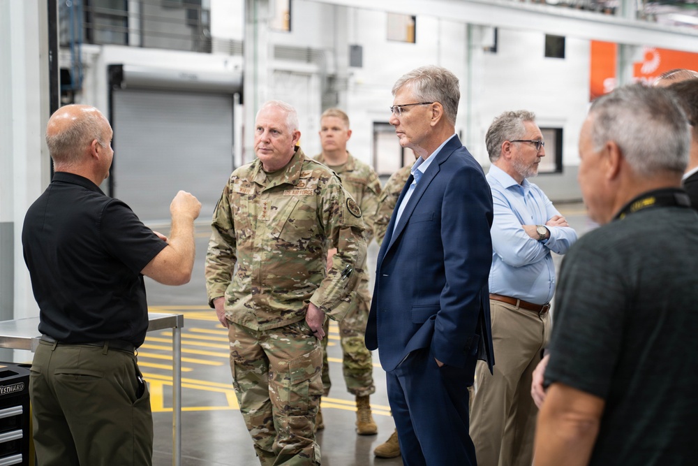 A group of individuals including US Air Force military personnel in OCP (Operational Camouflage Pattern) uniforms and civilians in business attire are gathered inside what appears to be an industrial or manufacturing facility. A man in a black polo shirt appears to be briefing or presenting to the group, gesturing with his hands. The setting suggests an official facility tour or inspection visit.