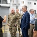 A group of individuals including US Air Force military personnel in OCP (Operational Camouflage Pattern) uniforms and civilians in business attire are gathered inside what appears to be an industrial or manufacturing facility. A man in a black polo shirt appears to be briefing or presenting to the group, gesturing with his hands. The setting suggests an official facility tour or inspection visit.
