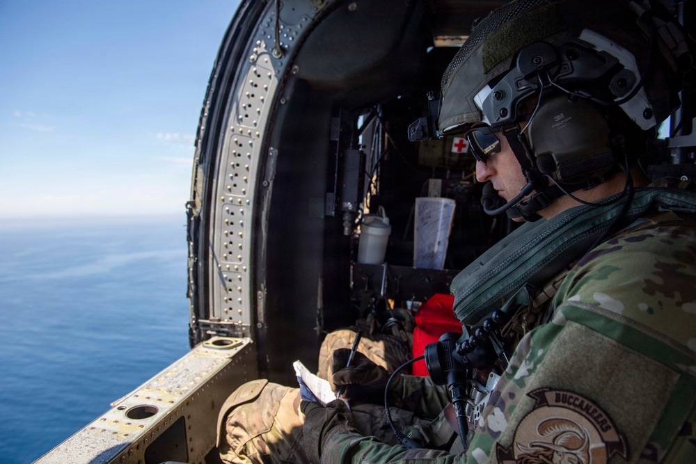 A military aviator or crew member is seated inside an open-door helicopter flying over a body of water, reviewing what appears to be a document or map. The individual is wearing full flight gear including a tactical helmet with communications equipment, camouflage uniform, and dark sunglasses. A red cross symbol is visible on equipment in the background, suggesting a medevac or search-and-rescue role.
