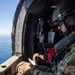 A military aviator or crew member is seated inside an open-door helicopter flying over a body of water, reviewing what appears to be a document or map. The individual is wearing full flight gear including a tactical helmet with communications equipment, camouflage uniform, and dark sunglasses. A red cross symbol is visible on equipment in the background, suggesting a medevac or search-and-rescue role.