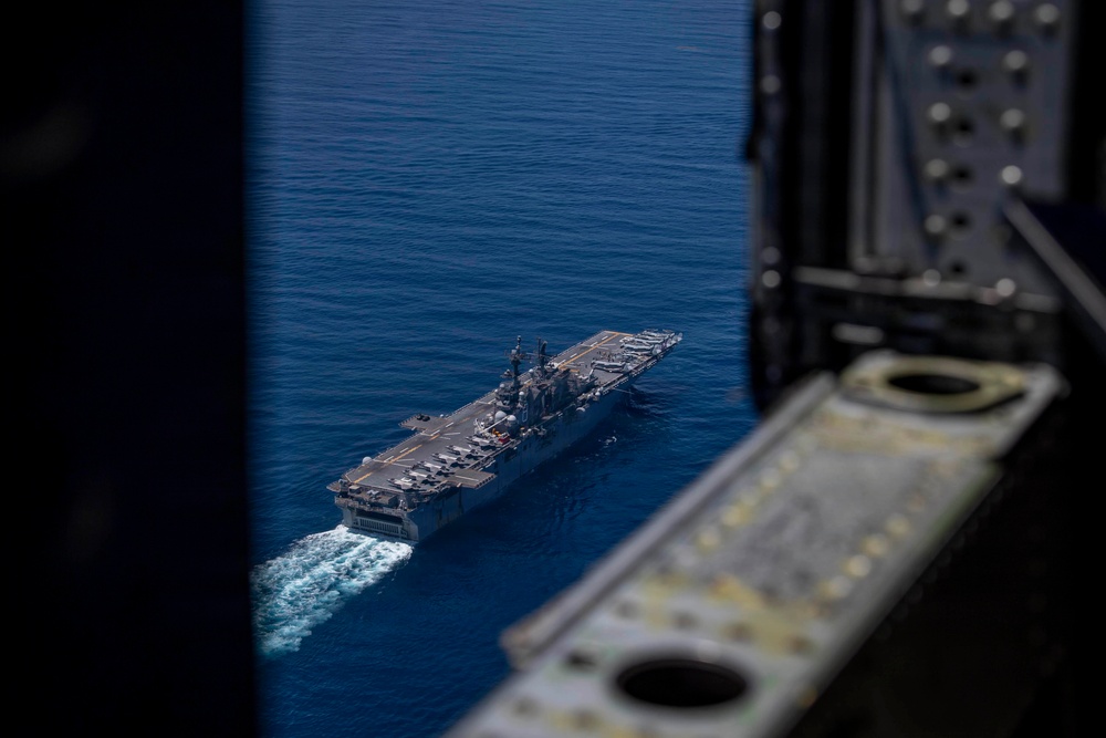 An aerial photograph taken from inside a military aircraft, looking through an open hatch or ramp at a large US Navy amphibious assault ship underway at sea. The flat-deck vessel, consistent with a Wasp-class or America-class LHA/LHD, is sailing through deep blue open ocean with a visible white wake. Multiple aircraft and equipment are visible on the flight deck.