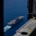 An aerial photograph taken from inside a military aircraft, looking through an open hatch or ramp at a large US Navy amphibious assault ship underway at sea. The flat-deck vessel, consistent with a Wasp-class or America-class LHA/LHD, is sailing through deep blue open ocean with a visible white wake. Multiple aircraft and equipment are visible on the flight deck.