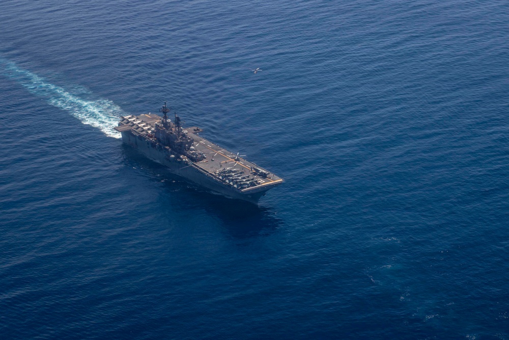 An aerial photograph of a large US Navy amphibious assault ship underway in open blue ocean waters. The flight deck is populated with multiple rotary-wing and fixed-wing aircraft, and the vessel is leaving a visible white wake behind it. A single aircraft appears to be airborne in the upper portion of the image.